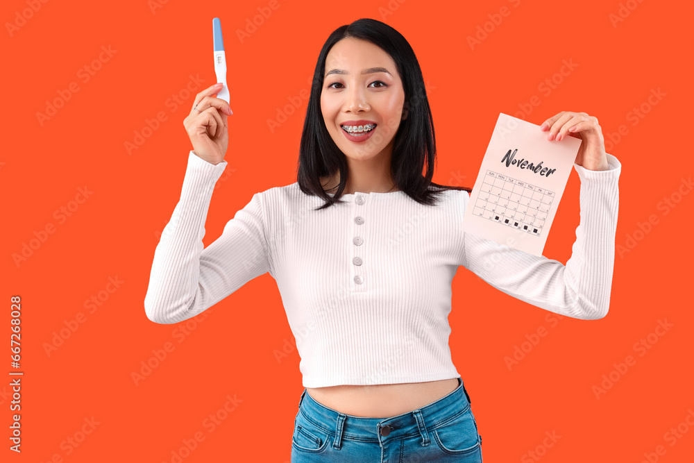 Young Asian woman with pregnancy test and calendar on orange background