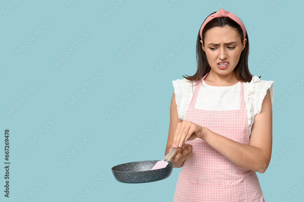 Portrait of annoyed young housewife in apron with frying pan and spatula on blue background