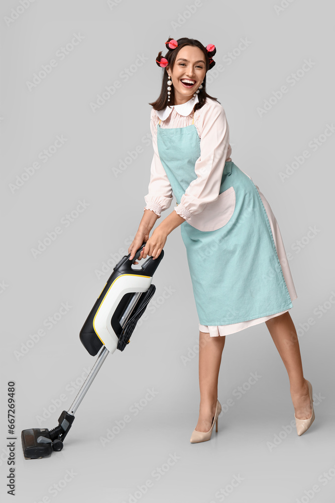 Portrait of happy young housewife in apron with vacuum cleaner on grey background