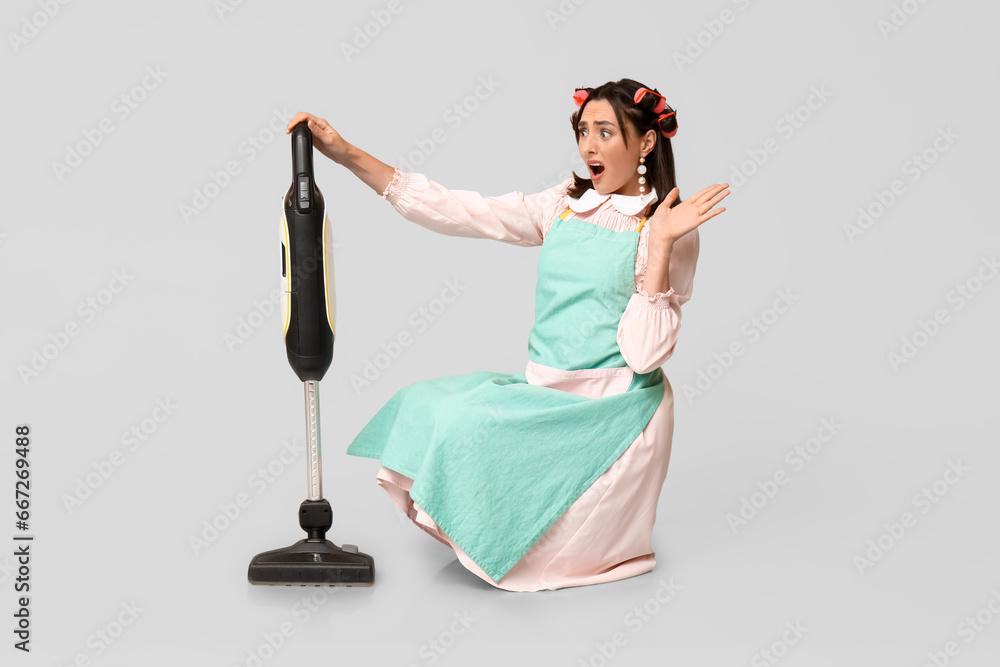 Portrait of shocked young housewife in apron with vacuum cleaner on grey background