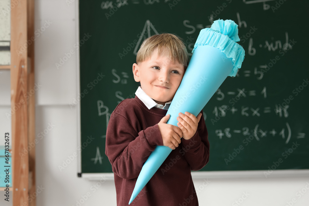 Happy little boy with blue school cone in classroom near blackboard