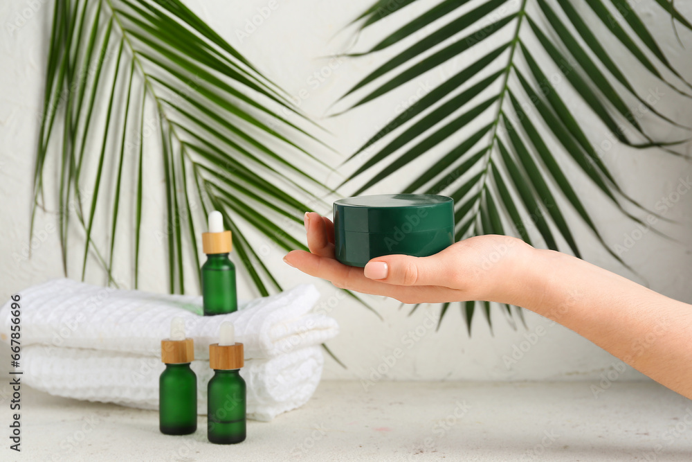 Female hand with cosmetic products, clean towels and palm leaves on light background