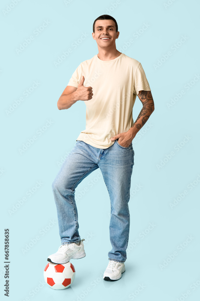 Handsome young man with soccer ball showing thumb-up gesture on blue background