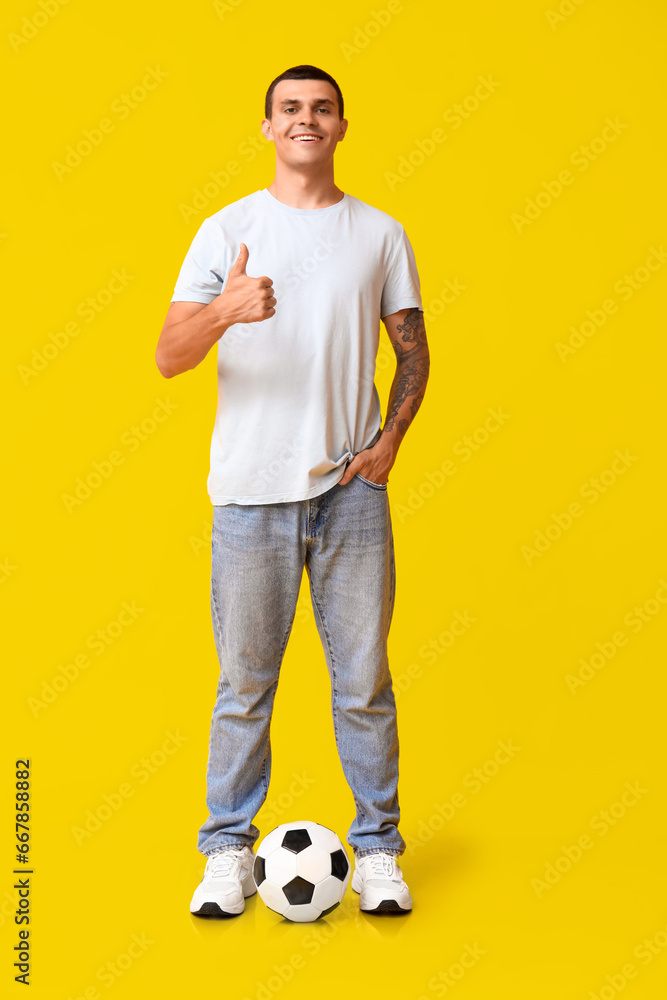 Handsome young man with soccer ball showing thumb-up gesture on yellow background