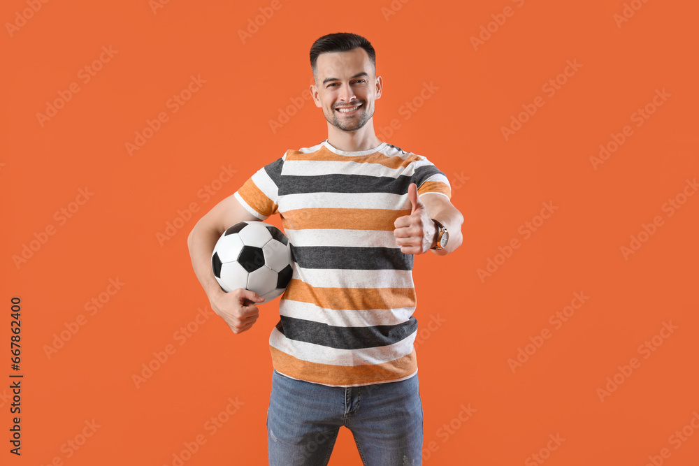 Happy young man with soccer ball showing thumb-up on orange background