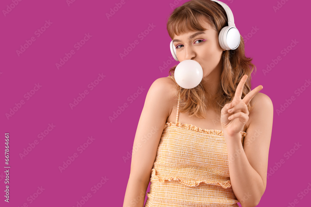 Young woman in headphones with chewing gum showing victory gesture on purple background