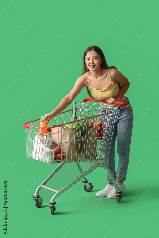 Young Asian woman with shopping cart and bags full of fresh food on green background