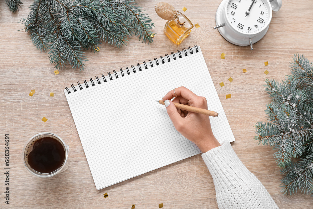 Woman writing in notebook with Christmas branches, coffee and perfume on light wooden background