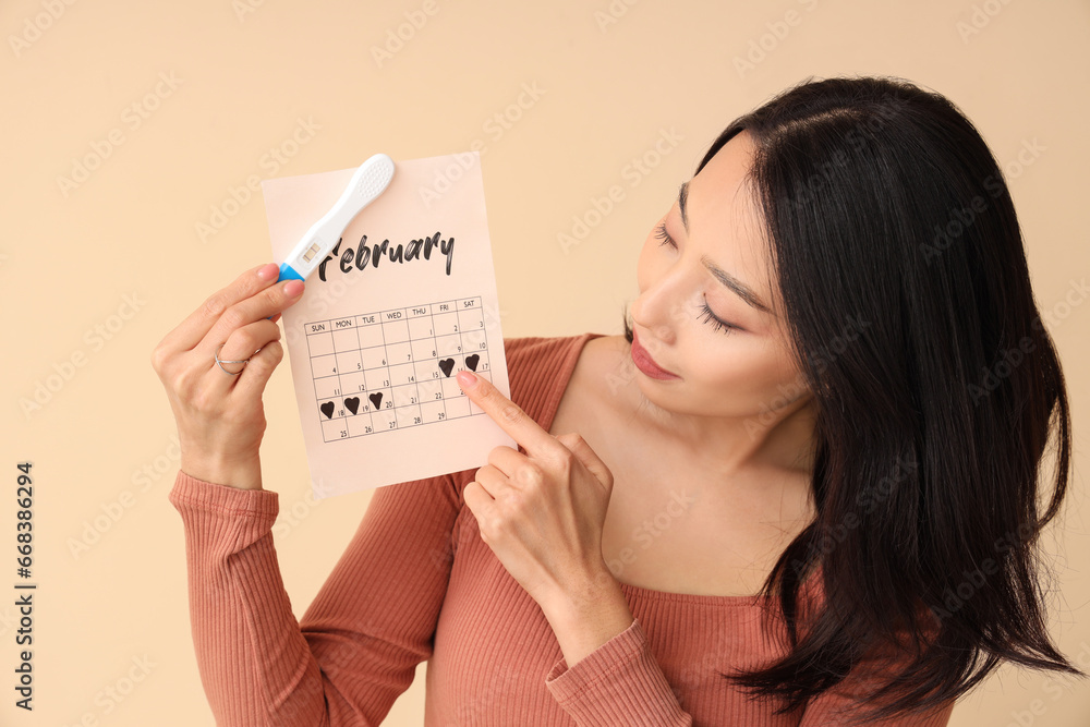 Young Asian woman with pregnancy test and calendar on beige background