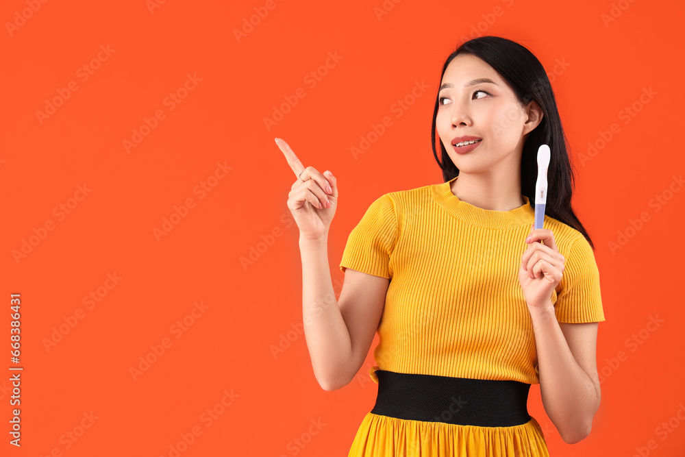 Young Asian woman with pregnancy test pointing at something on orange background