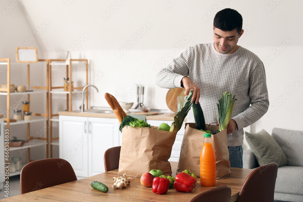 Young man taking fresh vegetables from grocery bags at table in kitchen