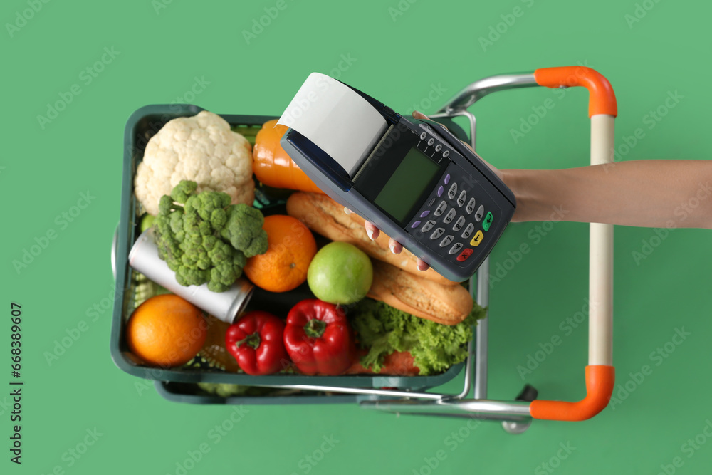 Shopping cart full of food and female hand with payment terminal on green background