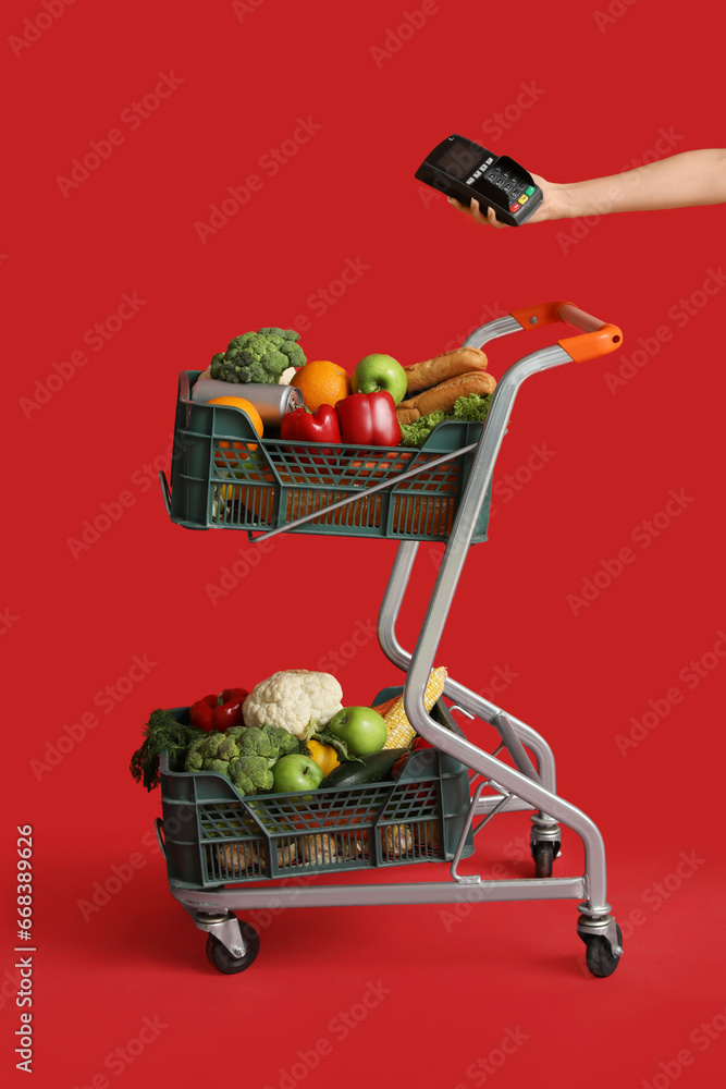 Shopping cart full of food and female hand with payment terminal on red background