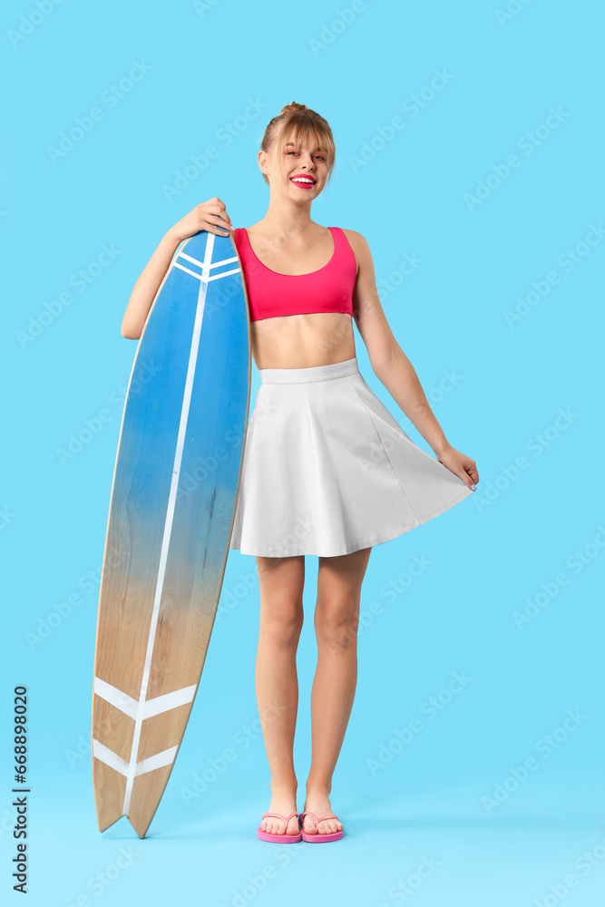 Young woman dressed as doll with surfboard on blue background