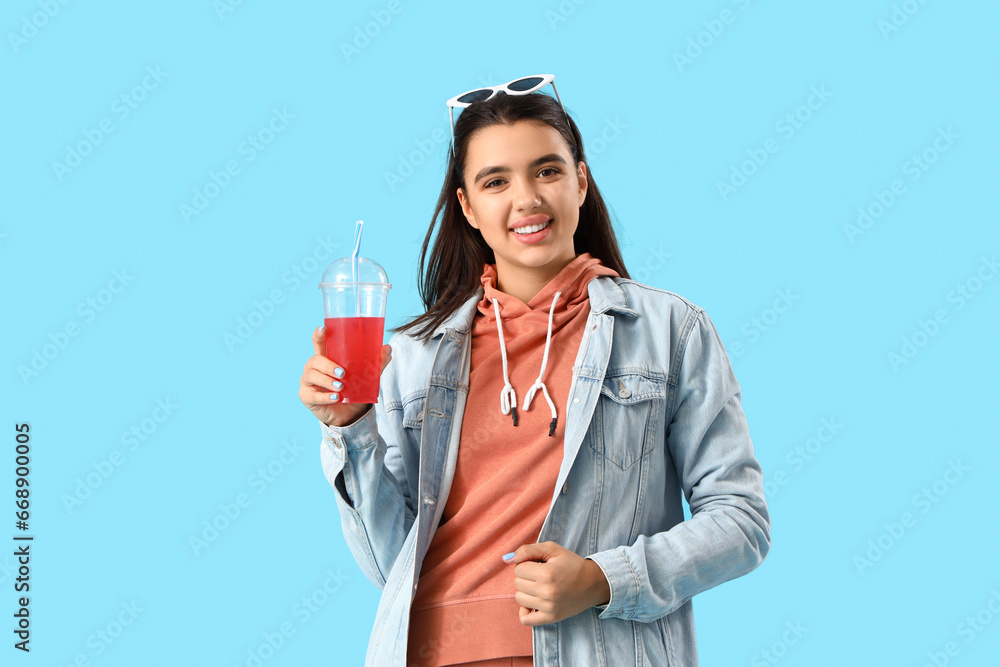 Beautiful young woman in stylish hoodie with cup of lemonade on blue background