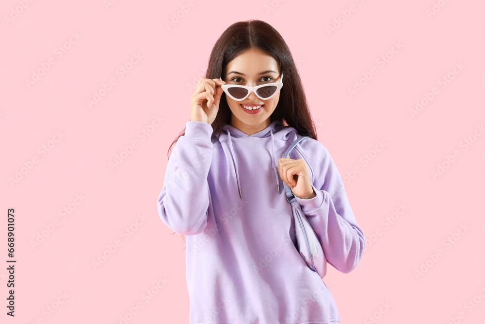 Beautiful young Asian woman in stylish hoodie and sunglasses on pink background