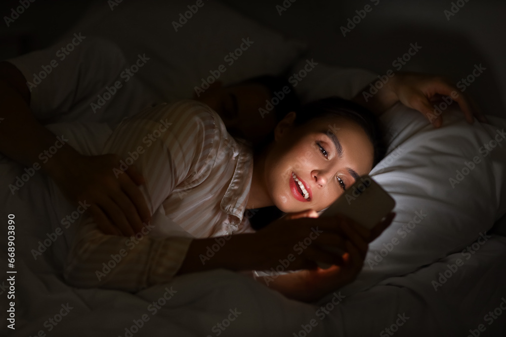 Young woman using mobile phone and her sleeping husband in bedroom at night