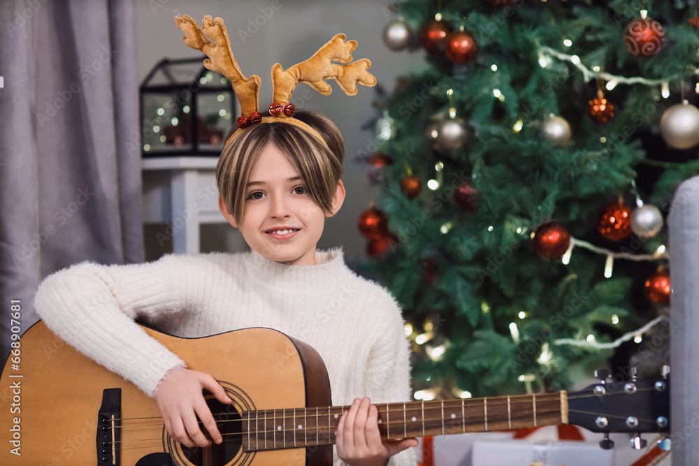 Cute little boy in deer horns playing guitar at home on Christmas eve