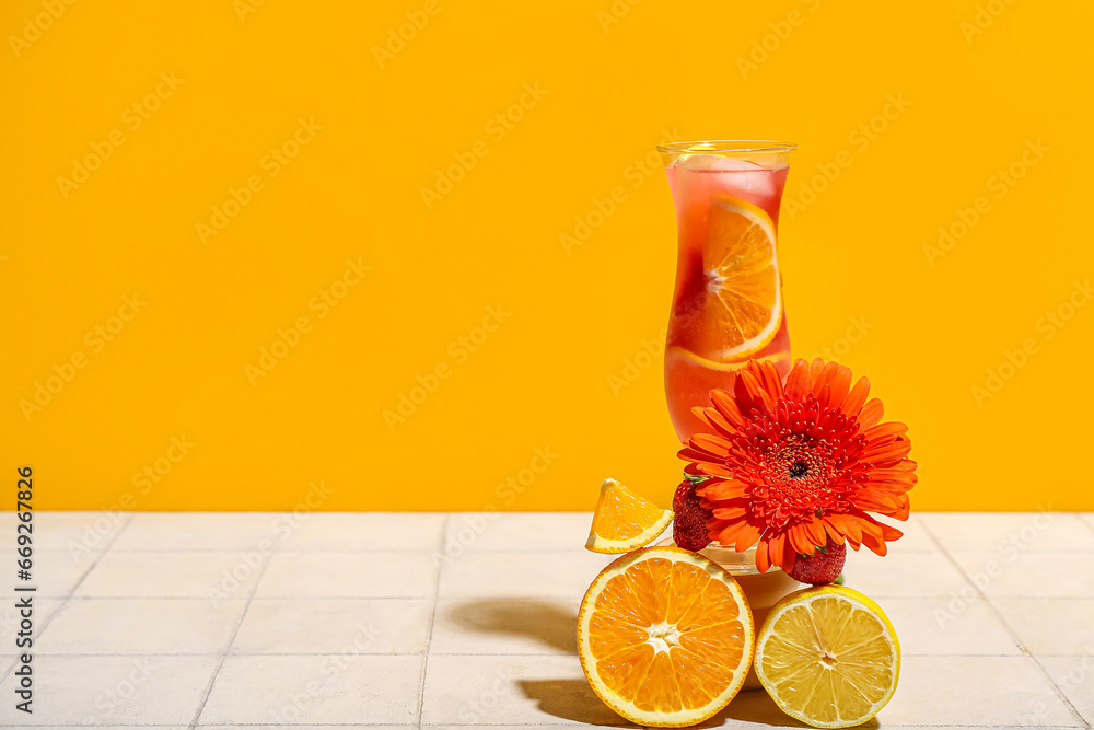 Glass of citrus cocktail with strawberries, lemon, orange and flower on white tile near yellow wall