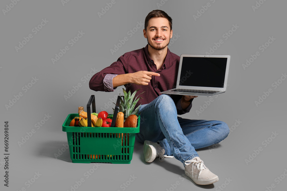 Young man with shopping basket pointing at laptop on grey background