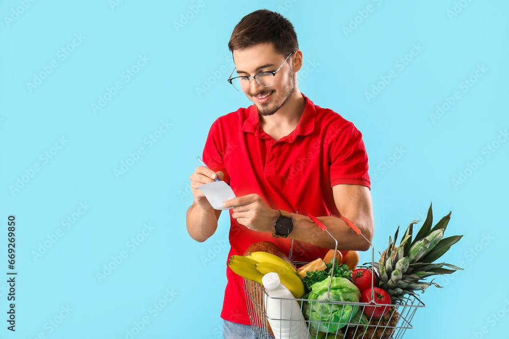 Young man with shopping basket checking grocery list on blue background