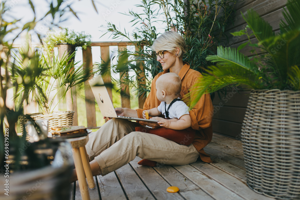 Mother holding baby while working on laptop in garden. Businesswoman working remotely from outdoor home office and taking care of little son. Life work balance with kid.