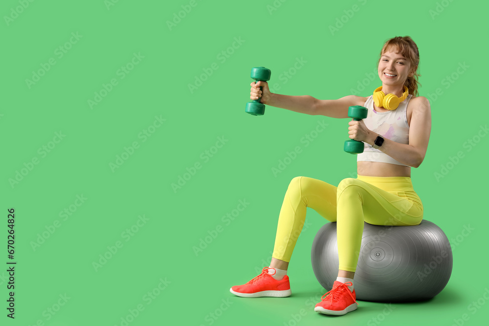 Young sporty woman with headphones and dumbbells sitting on fitness ball against green background