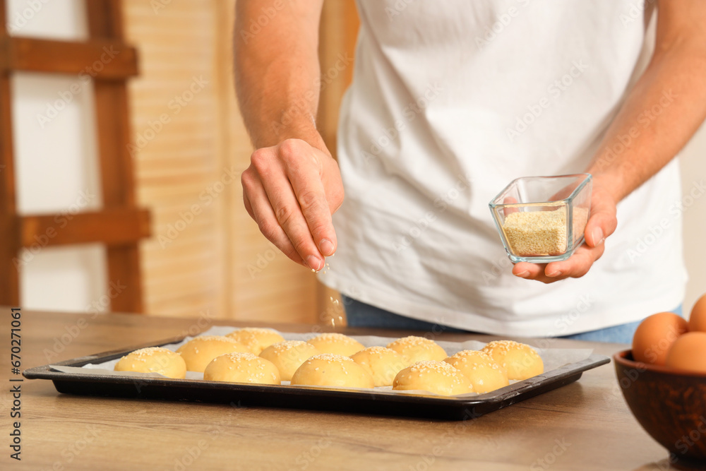 Handsome young man sprinkling raw buns with sesame seeds at table in bakery