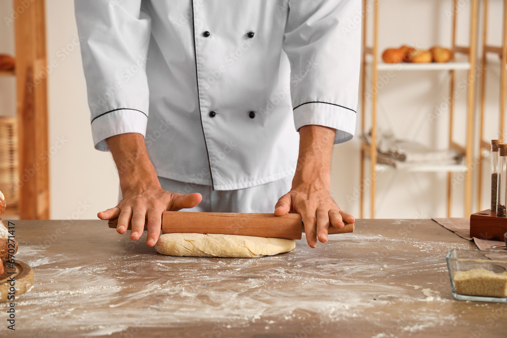 Young handsome chef rolling out dough on table in bakery, closeup