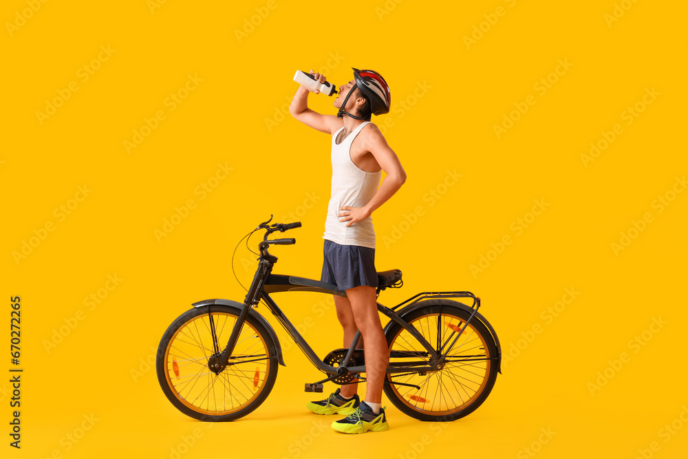 Young male cyclist with bicycle drinking water on yellow background