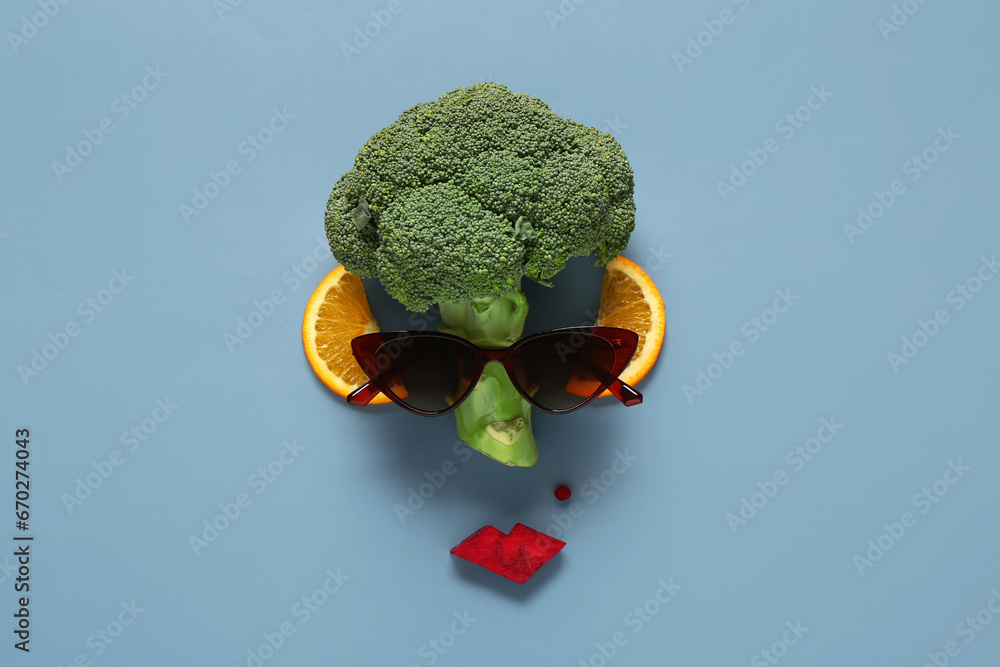 Funny face made of fresh broccoli, orange slices and sunglasses on color background