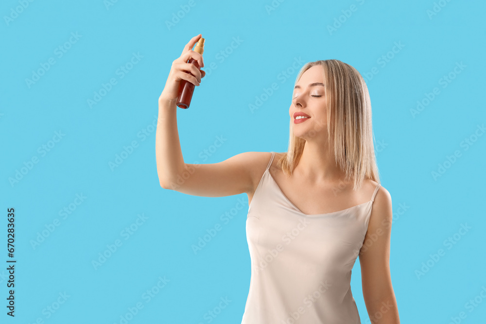 Young blonde woman with spray bottle of hair product on blue background