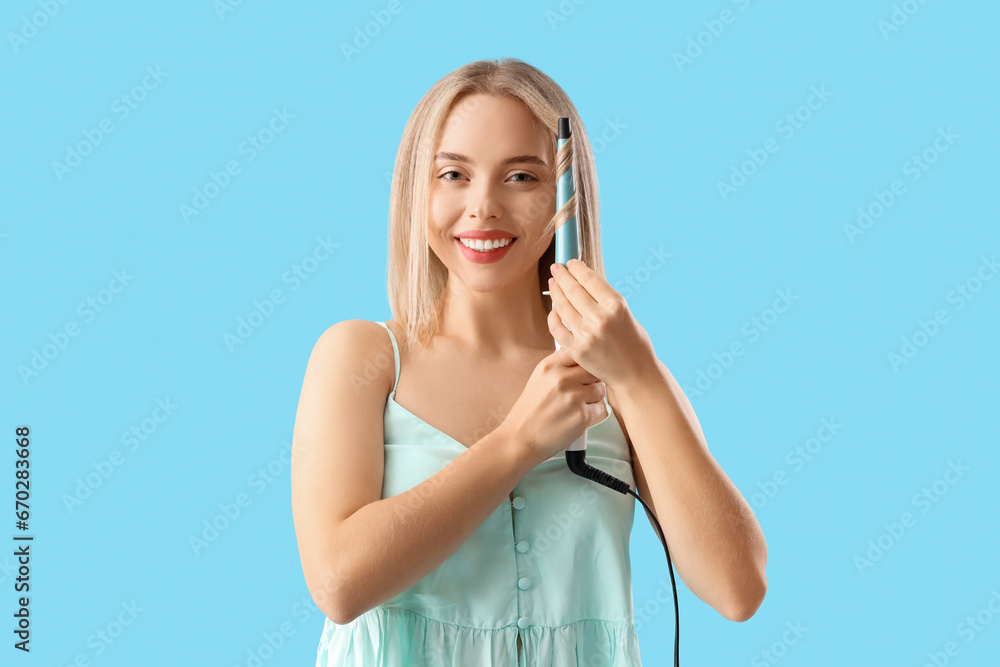 Young blonde woman curling hair on blue background