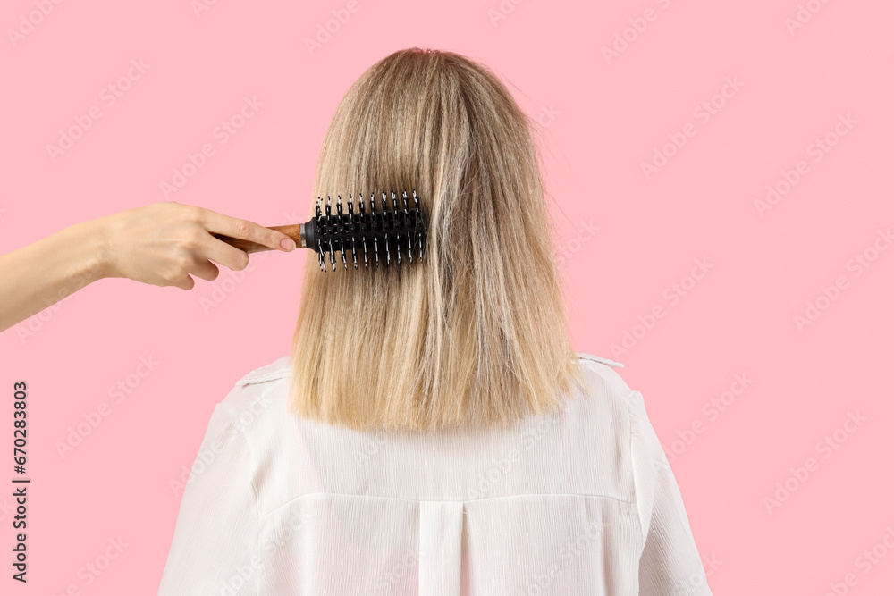 Young blonde woman brushing hair on pink background, back view