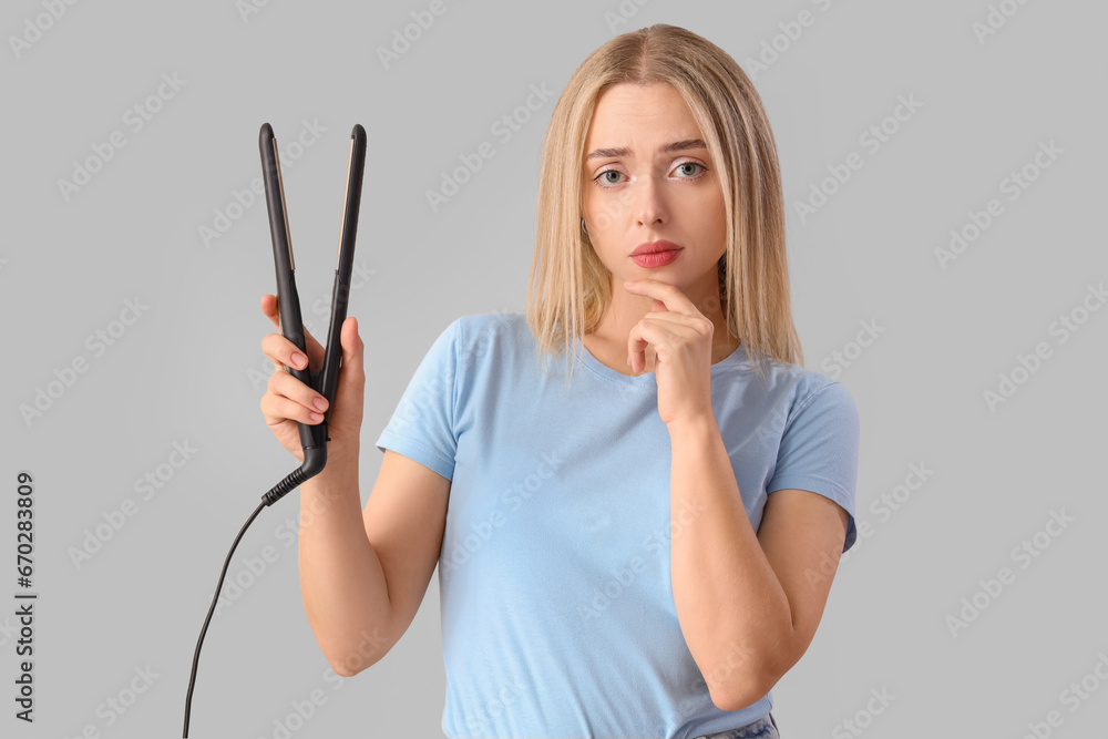 Thoughtful young blonde woman with straightener on light background