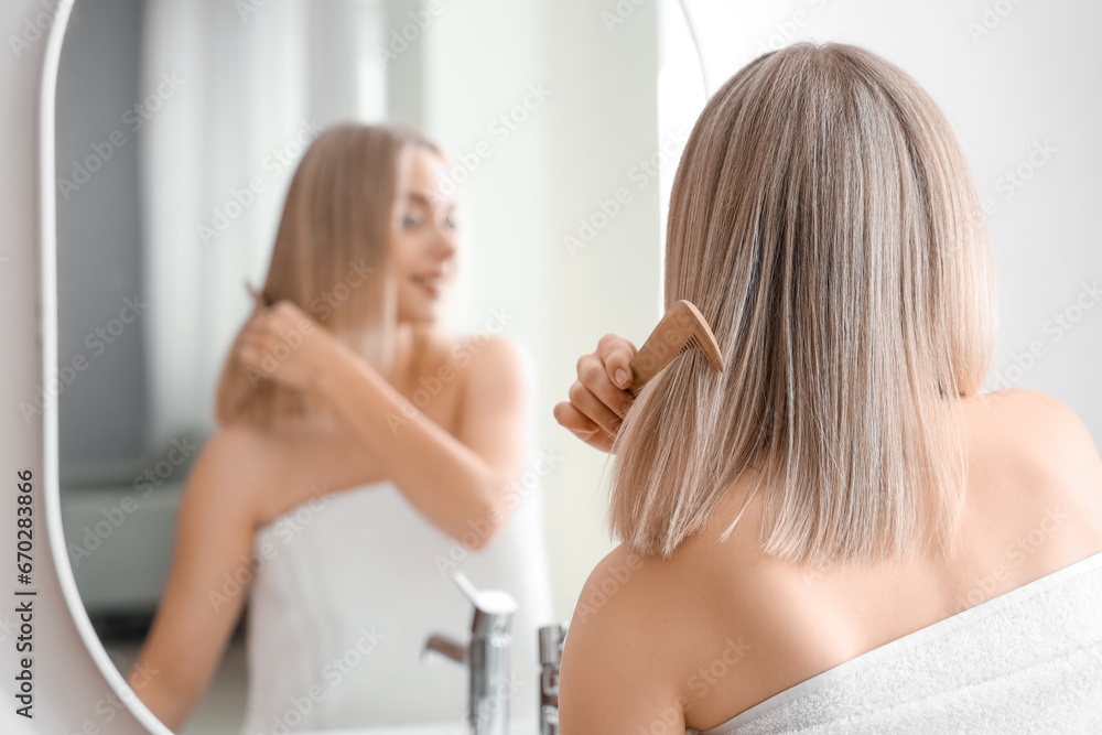 Young blonde woman combing hair near mirror in bathroom, back view