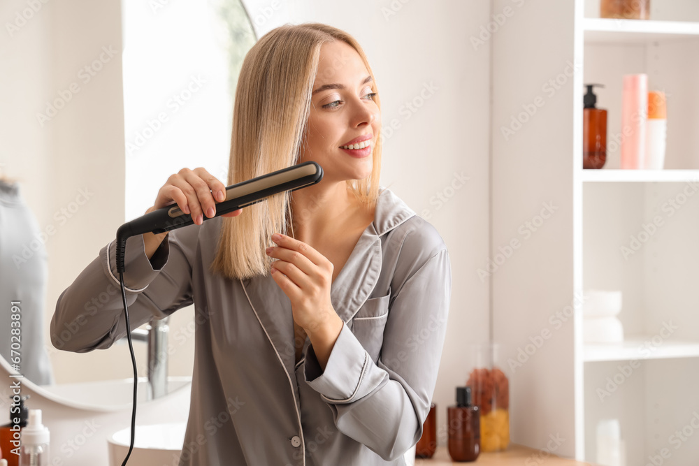 Young blonde woman straightening hair in bathroom