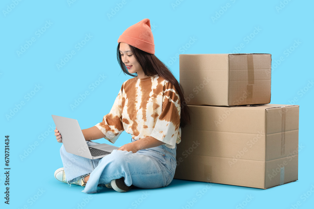 Young Asian woman with laptop and parcels on blue background