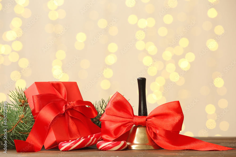 Christmas bell with gift box and candy cane on wooden table against blurred lights