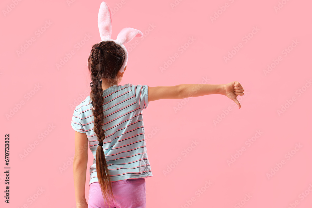 Little girl in bunny ears showing dislike on pink background, back view