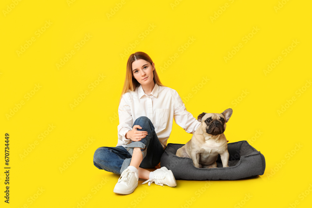 Beautiful young woman and cute pug dog sitting on pet bed against yellow background