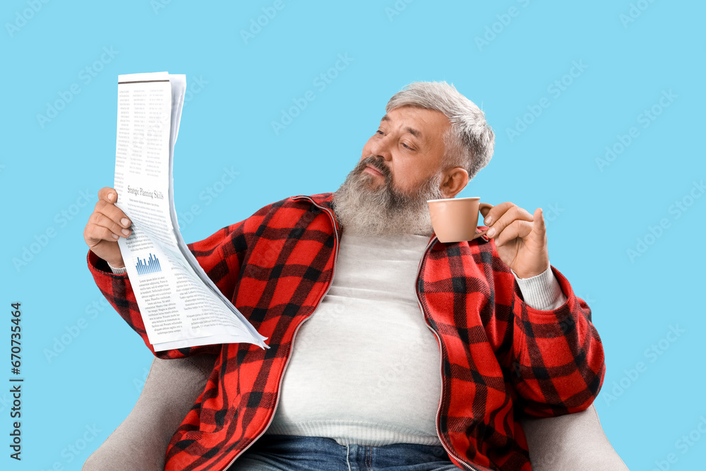 Mature man with cup of coffee and newspaper sitting on armchair against blue background