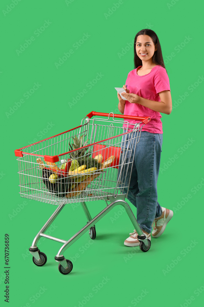 Young woman with shopping cart checking grocery list on green background