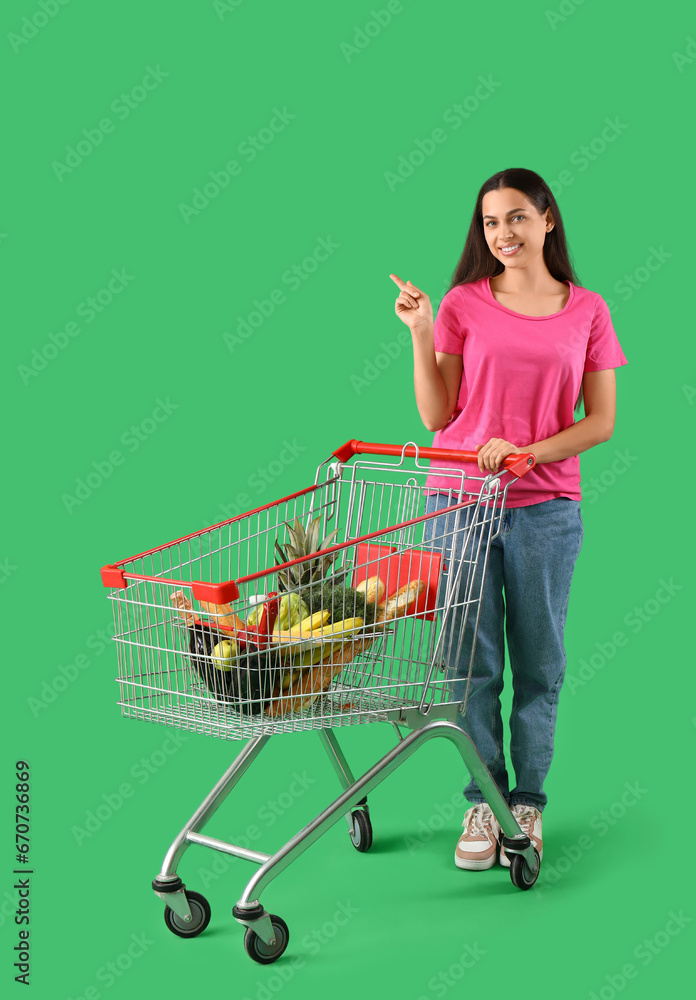 Young woman with shopping cart pointing at something on green background