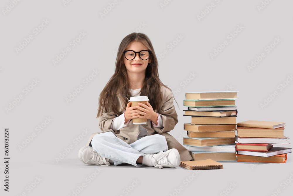 Little girl with cup of cocoa and books sitting on light background