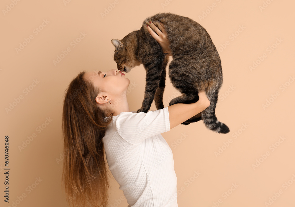 Pretty young woman kissing cute tabby cat on beige background