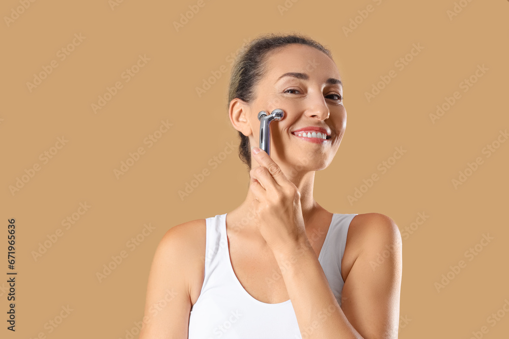 Beautiful mature woman with facial massage roller on beige background