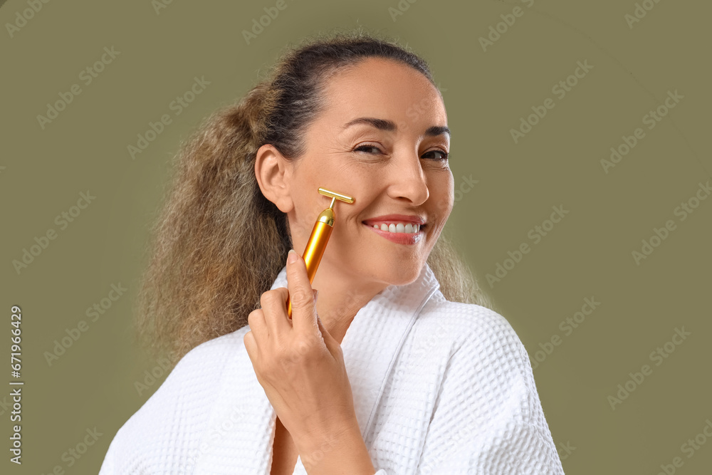 Beautiful mature woman using face massage roller on khaki background