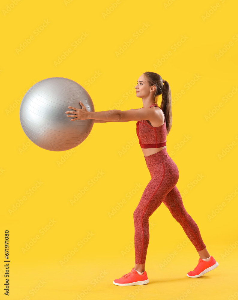 Sporty young woman training with fitball on yellow background