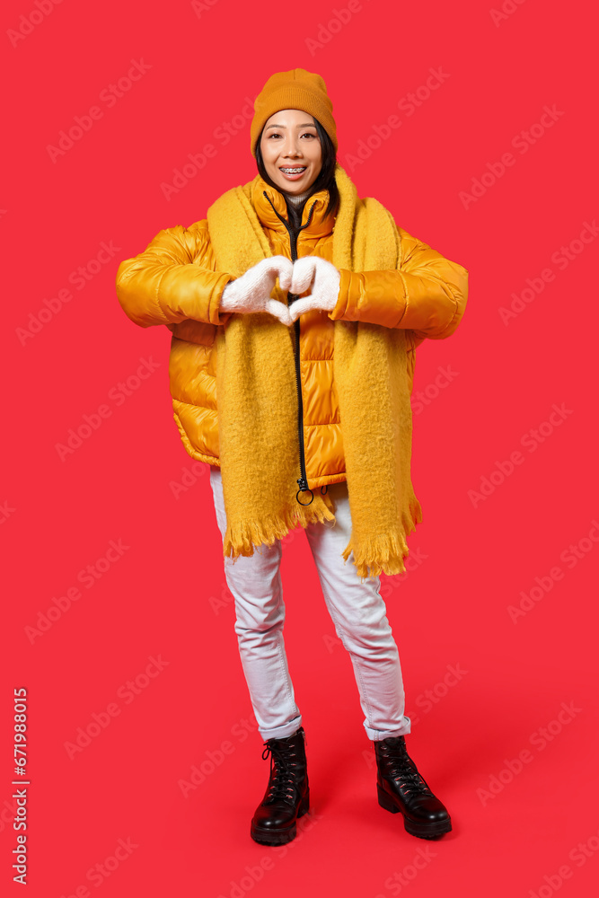 Young Asian woman in winter clothes making heart gesture on red background